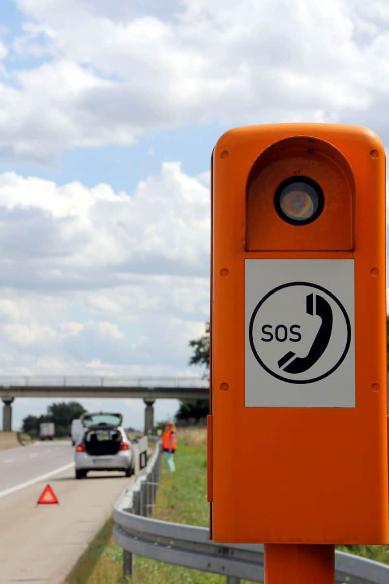 orange SOS phone box on the side of the road with a broken down car in the distance